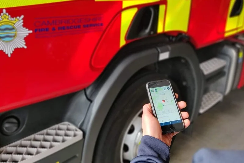 Person holding a smartphone showing a map application in front of a Cambridgeshire Fire & Rescue Service truck.
