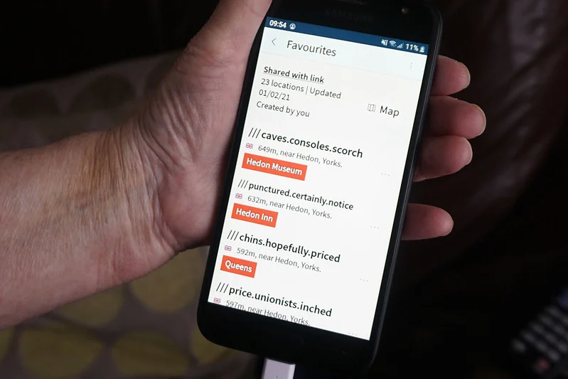 Hand holding a smartphone showing a list of favourite locations with geocodes, including Hedon Museum, Hedon Inn, and Queens.