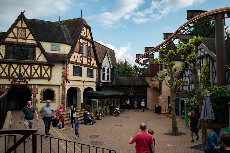 People walking and sitting in a medieval-themed amusement park area with a roller coaster track overhead and a gift shop on the left side.