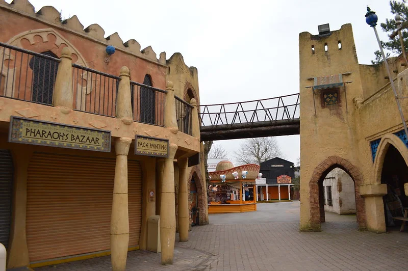 Desert-themed amusement park area with buildings featuring signage for "Pharaoh's Bazaar" and "Face Painting," and an arched pedestrian bridge above.