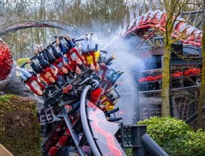People riding an inverted roller coaster with red and black tracks, surrounded by trees and mist.