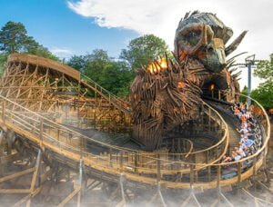 Wooden roller coaster with a large, intricate wooden structure resembling a mythical creature's head, surrounded by trees and a partly cloudy sky.