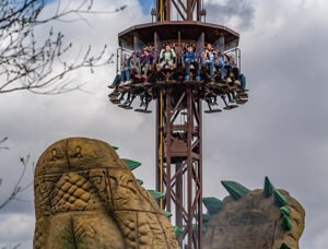 People riding a drop tower amusement ride with a dinosaur-themed structure in the foreground.