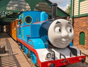 Blue and red train with a smiling face at a station platform with green-roofed shelter.