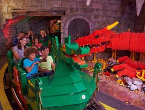 People riding a dragon-themed amusement park ride, passing by a large dragon sculpture made of Lego bricks.