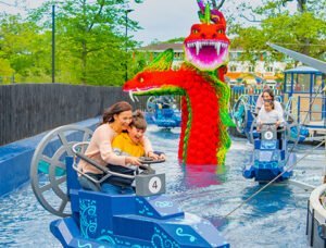 A woman and child ride a boat shaped like a ship amid a water attraction with a large colourful dragon sculpture in the background.