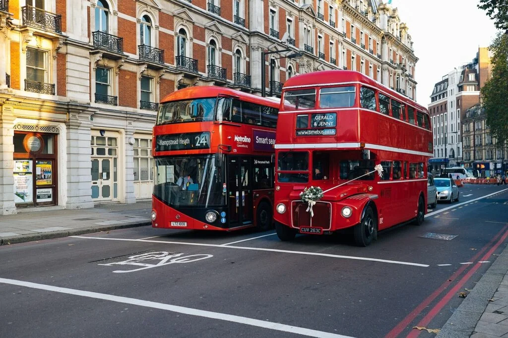Two red double-decker buses on a street in London, with a traditional Routemaster bus decorated for a wedding beside a modern bus.