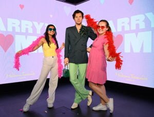 Two women posing with a wax figure of a man at Madame Tussauds London, with colourful feather boas and sunglasses against a backdrop that says "Harry" and "Madame Tussauds London".