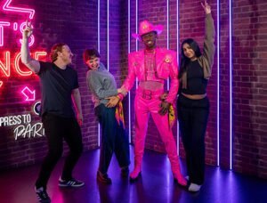 Four people posing in front of a neon-lit brick wall, one wearing a bright pink outfit with a matching cowboy hat.