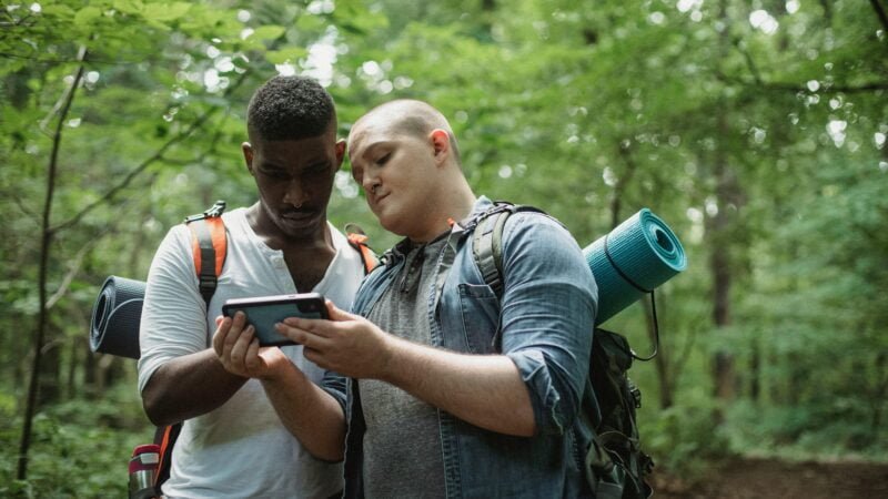 Two people with backpacks examining a mobile phone in a forest.