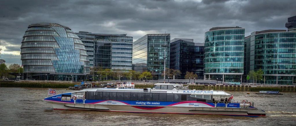 Modern buildings along the River Thames with a passenger ferry in the foreground.