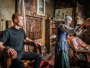 The London Dungeon 2 A young man is tied to a chair while a woman dressed in historical attire gestures dramatically in a dimly lit, medieval-themed room filled with old surgical instruments and anatomical drawings.