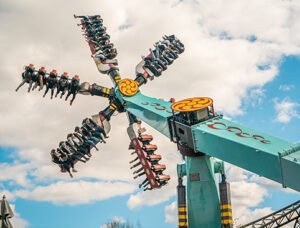 THORPE PARK Resort 3 People riding an amusement park thrill ride shaped like a large spinning windmill against a blue sky with clouds.