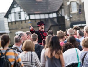 A Yeoman Warder in traditional uniform talking to a group of tourists outside historic buildings.