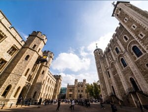 A view of the Tower of London with its towers and stone buildings under a partly cloudy sky, with people walking in the courtyard.