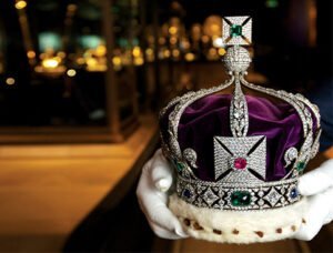A richly decorated crown adorned with diamonds, emeralds, and other gemstones, held by gloved hands.