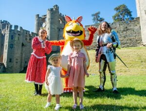 Two children pose with a knight, a person in a dragon costume, and a woman in a red and white historical dress in front of a medieval castle.