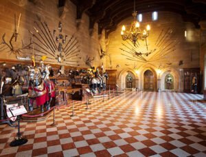 Grand hall with checkered floor, displaying suits of armour and various medieval weapons on the walls, illuminated by a central chandelier.