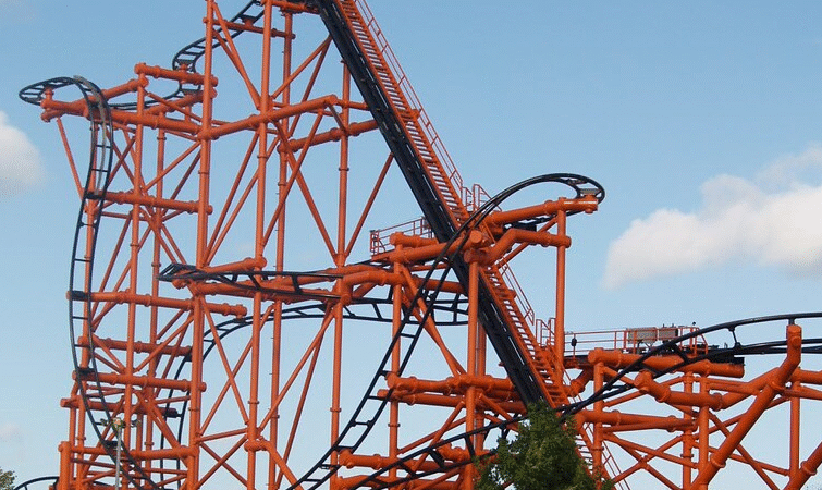 Tall roller coaster structure with bright orange supports and black tracks under a clear blue sky with a few clouds, situated near green grass and trees.